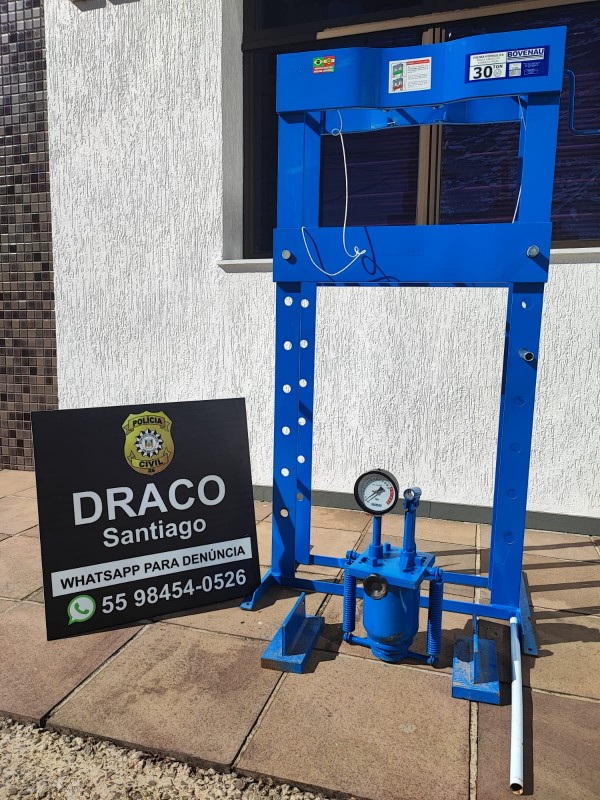Prensa hidráulica na cor azul, ao lado de placa da Draco de Santiago da Polícia Civil, com fundo em parede branca