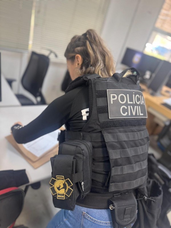 Policial mulher com coleta preto com escrita "Polícia Civil" folheando documentos sobre uma mesa