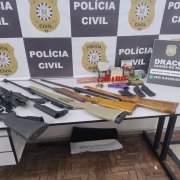 Diferentes armas de fogo e outros materiais como munições expostos em uma mesa com banner da Polícia Civil ao fundo e placa da Draco de Caxias do Sul ao lado