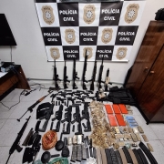 Armas e munições dispostos ao chão com banner da Polícia Civil ao fundo