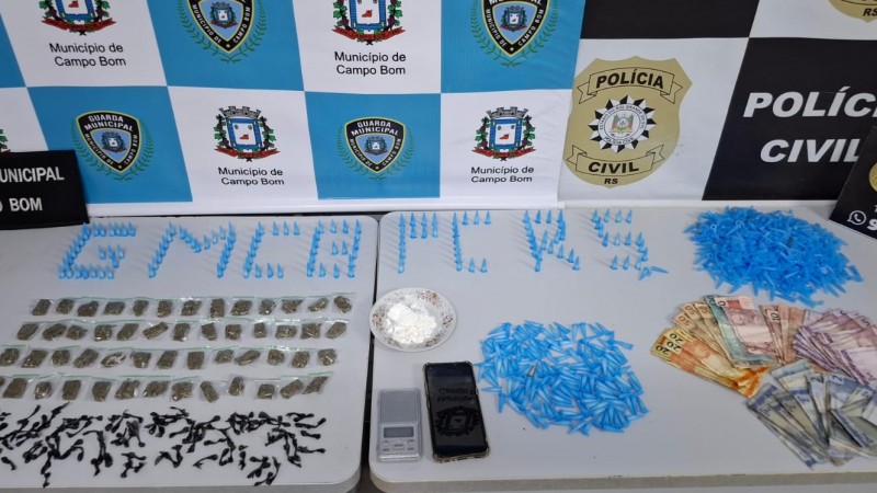 Pinos, entorpecentes, dinheiro e outros materiais apreendidos dispostos sobre uma mesa branca, com banners da Pol&iacute;cia Civil e Guarda Municipal de Campo Bom ao fundo