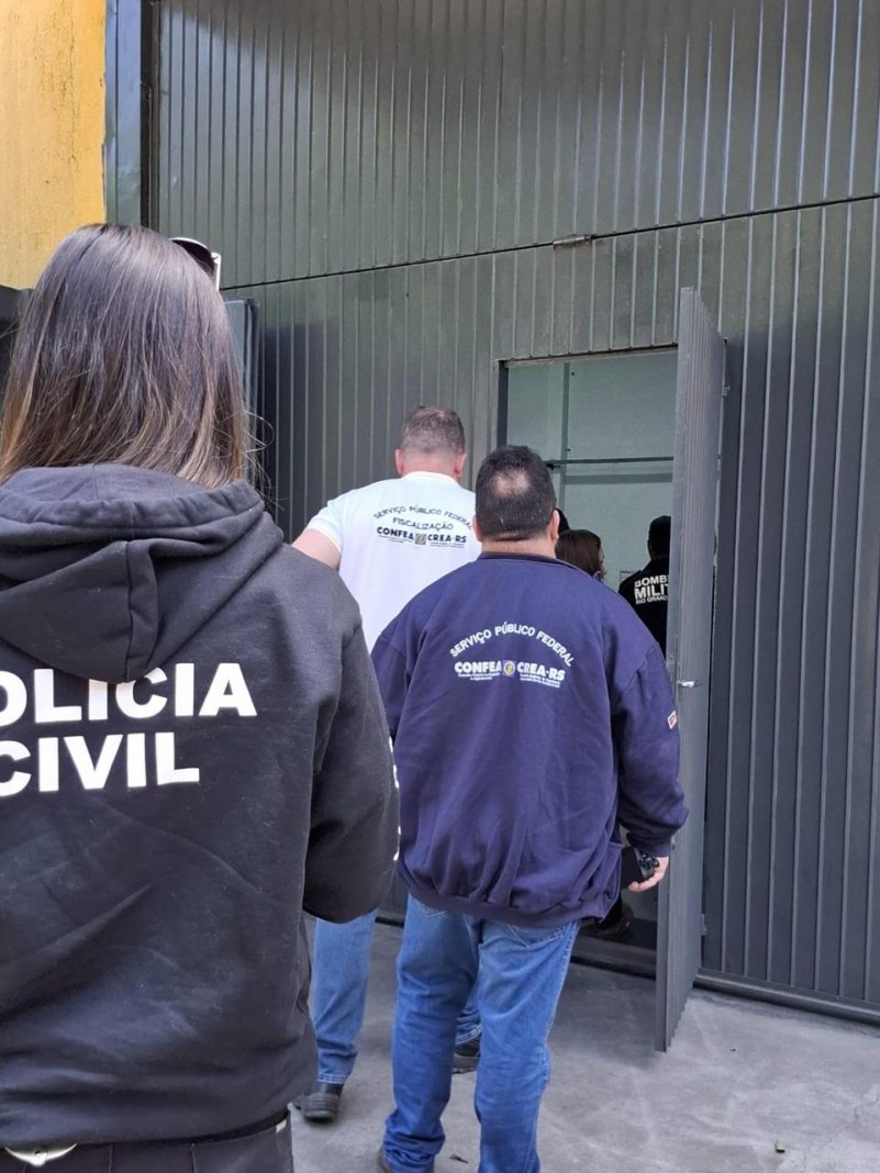 Agentes da Polícia Civil, Crea-RS, Corpo de Bombeiros e Prefeitura de Campo Bom de costas em fila entrando em uma porta de garagem grande e preta