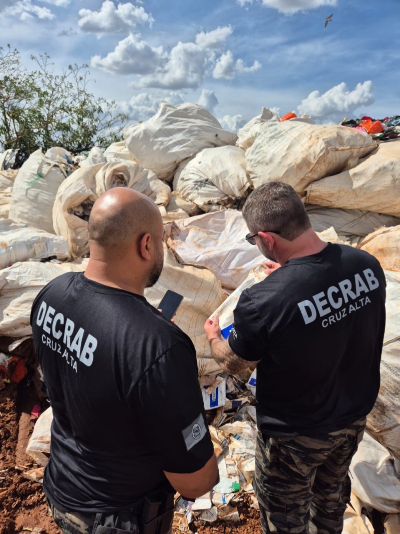 Dois homens de costas vestindo camisetas pretas com os inscritos: Decrab Cruz alta com diversos materiais ao fundo