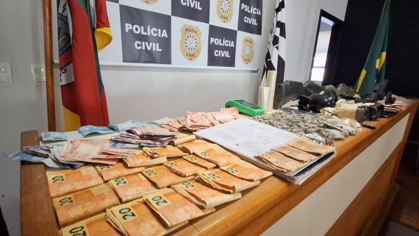 C&eacute;dulas de dinheiro, drogas e demais itens expostos em uma mesa, tendo o banner da Pol&iacute;cia Civil ao fundo