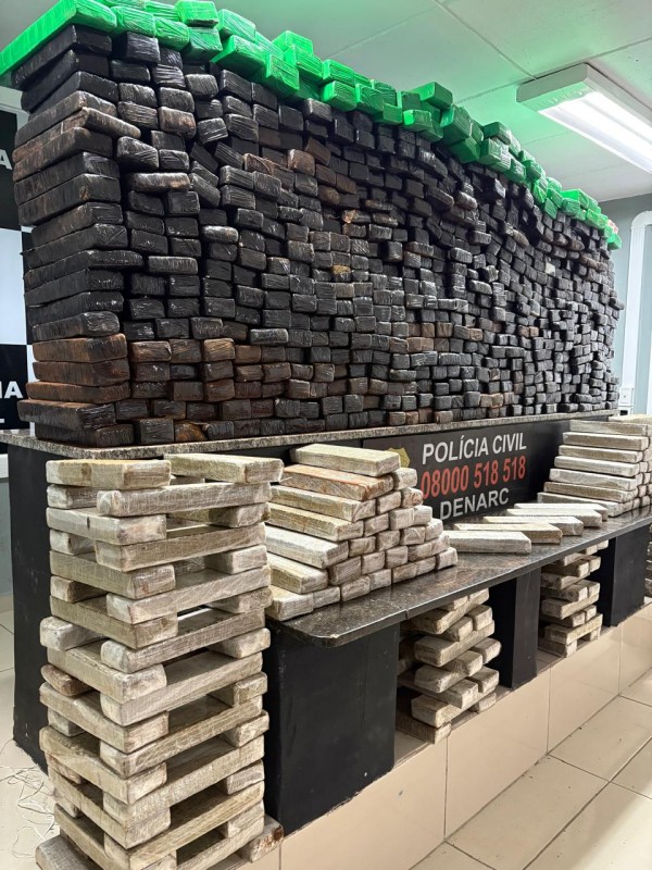 Diversos tabletes de drogas de cores claras e escuras dispostos em cima de mesa com dizeres Polícia Civil e denúncia 0800 518 518