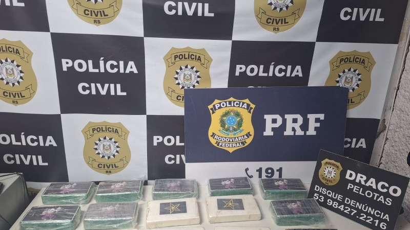 Tabletes brancos e esverdeados dispostos em cima de mesa com banner da Pol&iacute;cia Civil ao fundo e placas da Pol&iacute;cia Civil e PRF