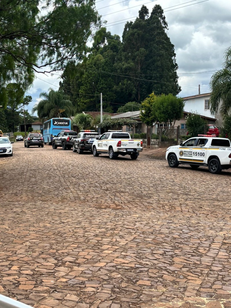 Veículos da Polícia Civil e Brigada Militar estacionados em rua de calçamento, com ônibus ao fundo