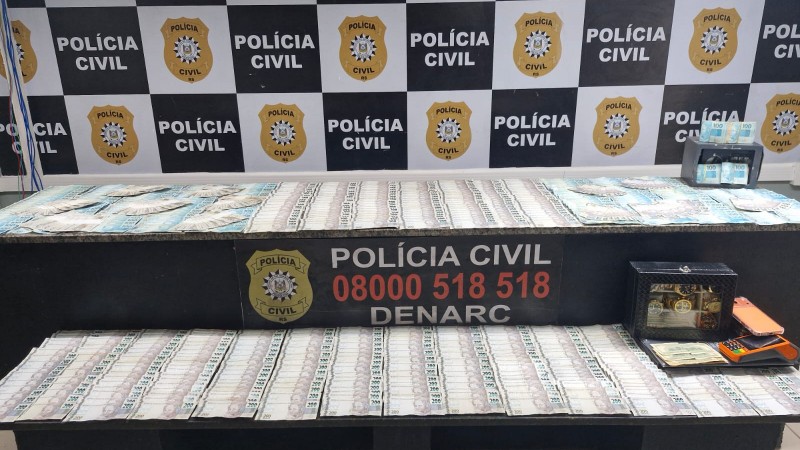 Diversos c&eacute;dulas de reais espalhadas sobre mesas e ao fundo banner da Pol&iacute;cia Civil