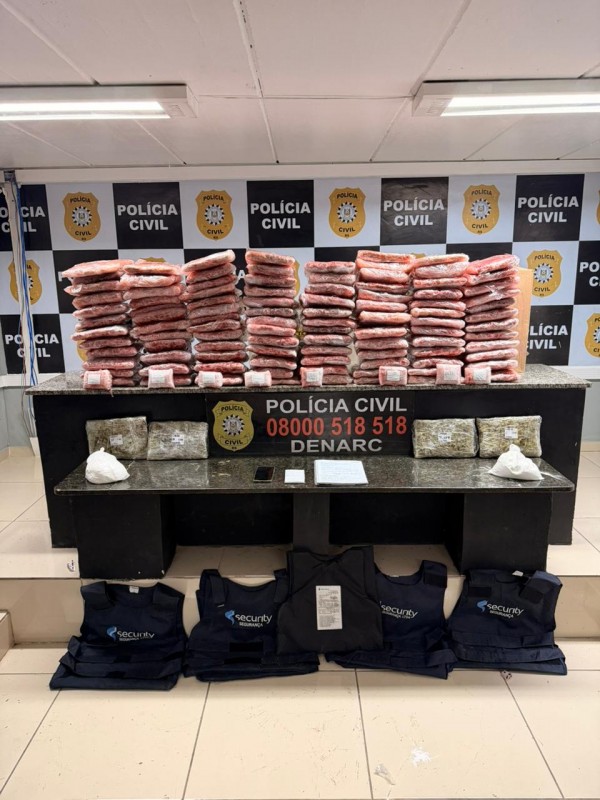 Pacotes de cor avermelhada dispostos em cima de mesa com banner da pol&iacute;cia civil ao fundo, al&eacute;m de coletes bal&iacute;sticos e outros materiais apreendidos