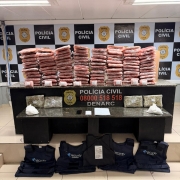 Pacotes de cor avermelhada dispostos em cima de mesa com banner da pol&iacute;cia civil ao fundo, al&eacute;m de coletes bal&iacute;sticos e outros materiais apreendidos