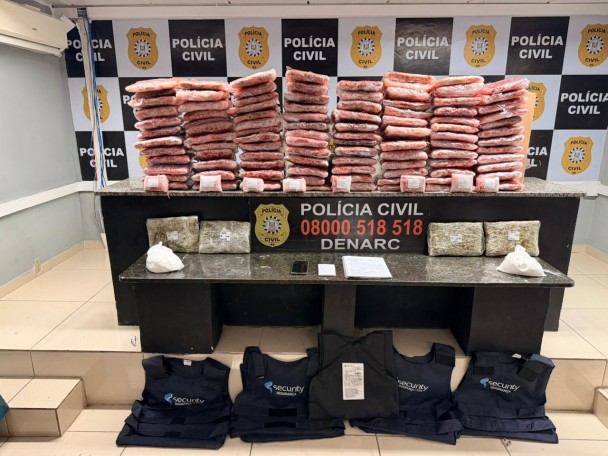 Pacotes de cor avermelhada dispostos em cima de mesa com banner da polícia civil ao fundo, além de coletes balísticos e outros materiais apreendidos