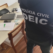 Policial de costas com dizeres Pol&iacute;cia Civil Deic no casaco e computadores, folhas e eletr&ocirc;nicos em mesa ao fundo