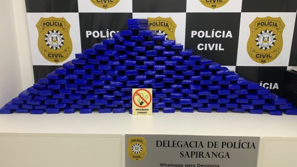 Drogas embaladas em envelopes azuis empilhados em formato de tri&acirc;ngulo em cima de uma mesa com a logo da pol&iacute;cia ao fundo