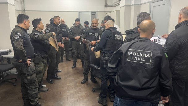 Diversos policiais homens vestidos com roupas e coletes da Pol&iacute;cia Civil, roupas e equipamentos majoritariamente pretos, dentro de uma sala com paredes brancas