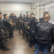 Diversos policiais homens vestidos com roupas e coletes da Pol&iacute;cia Civil, roupas e equipamentos majoritariamente pretos, dentro de uma sala com paredes brancas