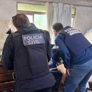 Policial civil mulher e perito criminal homem de costas observando o que deduz-se ser algum equipamento eletr&ocirc;nico, mas n&atilde;o vis&iacute;vel na imagem, dentro de uma esp&eacute;cie de sala ou quarto