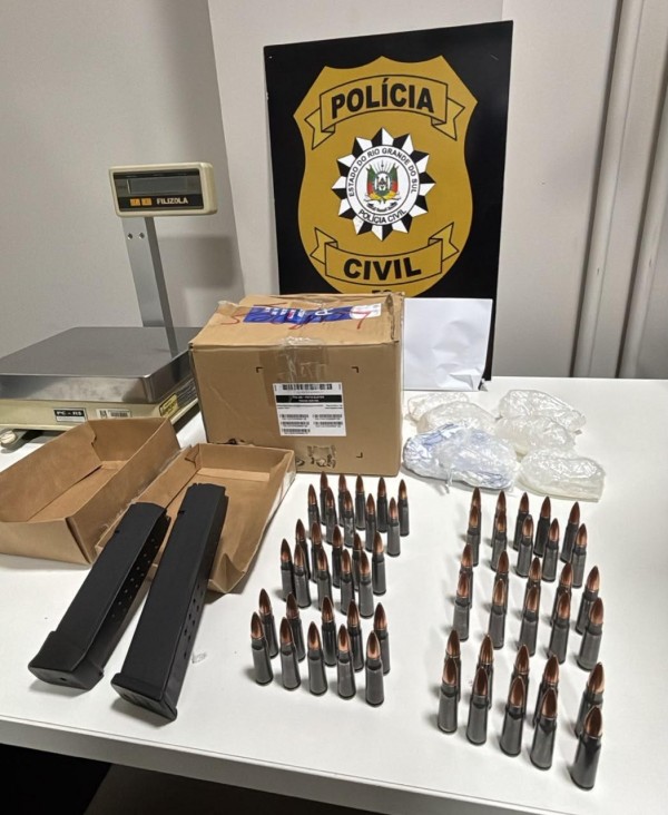 Cerca de 20 munições de fuzil de grosso calibre dispostas em cima de uma mesa, junto com caixas de papelão e ao fundo brasão da Polícia Civil