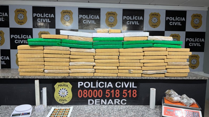 Diversos tabletes de drogas embalados em colora&ccedil;&atilde;o amarela e verde, al&eacute;m de outros materiais citados no texto da mat&eacute;ria expostos em bancadas com banner da Pol&iacute;cia Civil ao fundo